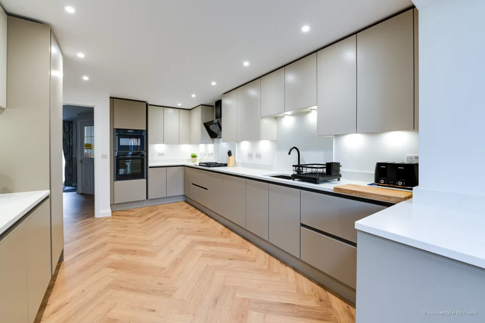 Wide pale grey handleless kitchen, pale worktop, black sink and tap, oven bank beyond.