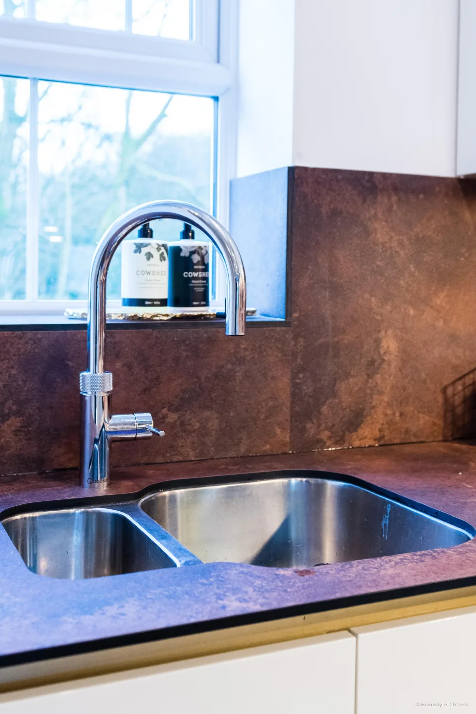 Stainless 1.5-bowl undermount sink and chrome tap in a copper-effect worktop, under a window.