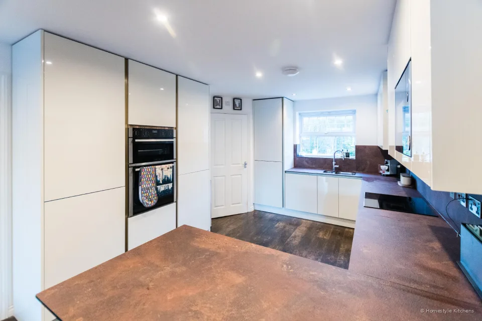 Gloss white kitchen from the doorway, oven stack to the left, window and copper-effect worktop to the right.
