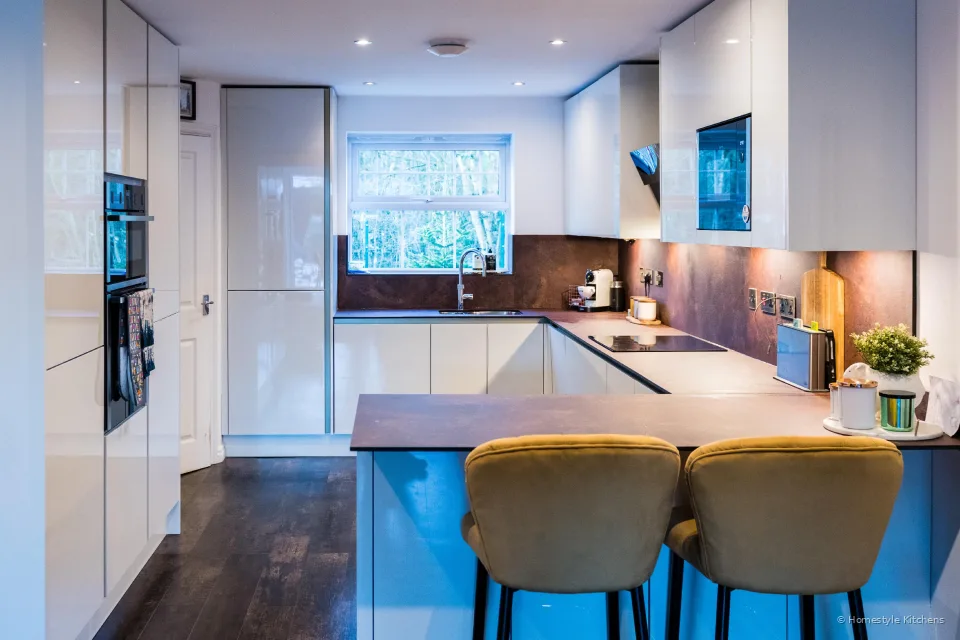 Gloss white galley kitchen looking down to a peninsula breakfast bar with mustard stools, copper-effect worktop.
