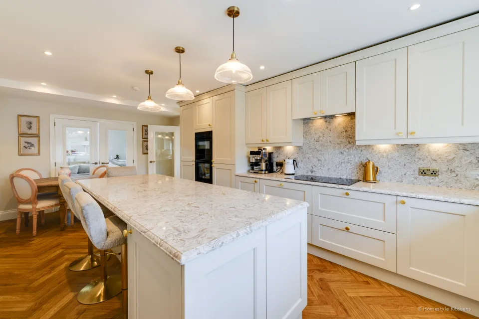 Cream painted kitchen with an island, three glass pendants, marbled quartz worktops and a herringbone floor.