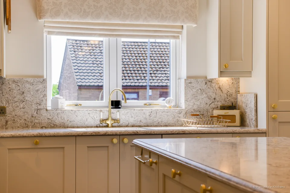Window over a marbled quartz worktop and splashback, roman blind and cream cabinets.