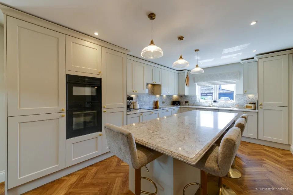 Cream painted kitchen island with glass pendants, tall oven housing and a window run beyond.