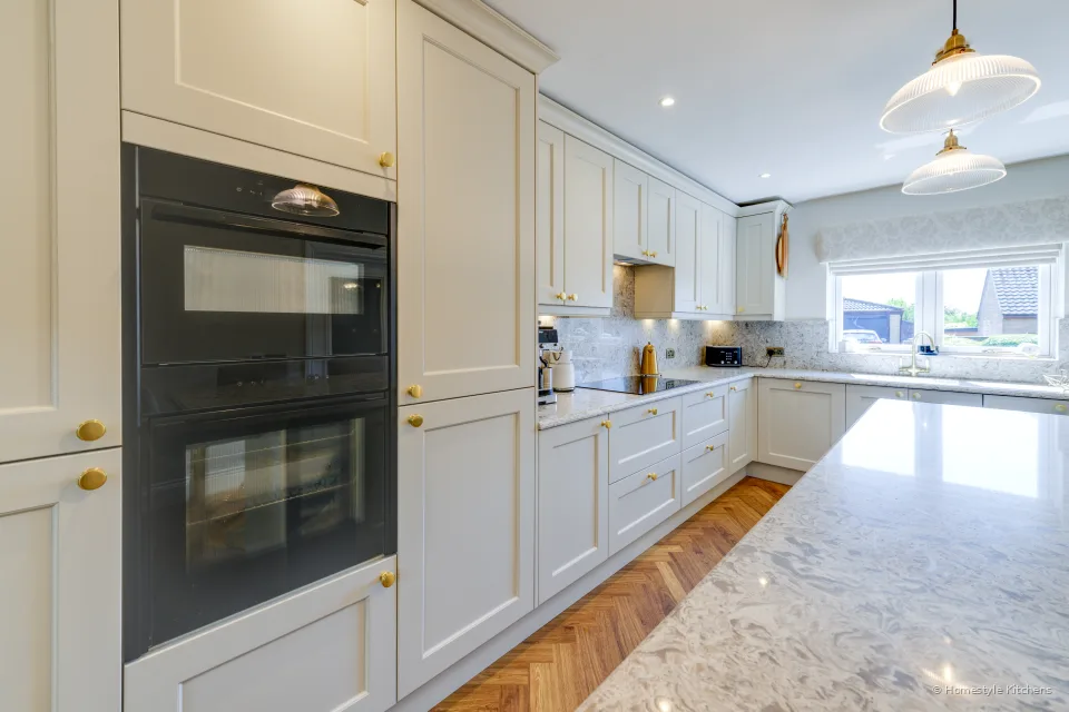 Tall cream cabinet housing black ovens, cream painted run down to a window, marbled quartz splashback.