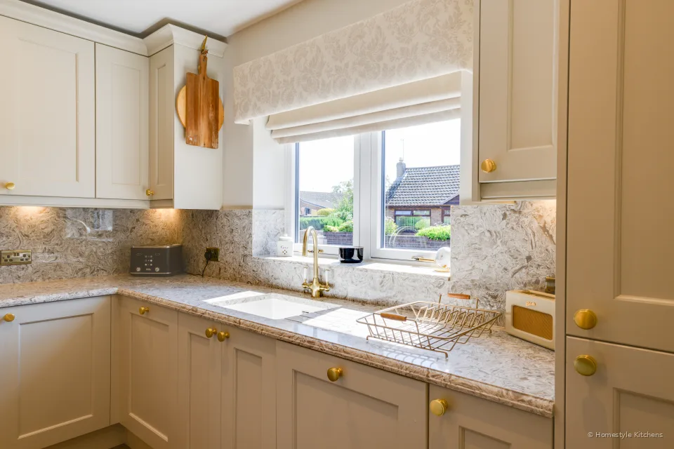 Cream painted corner run under a window, marbled quartz worktop and brass tap.