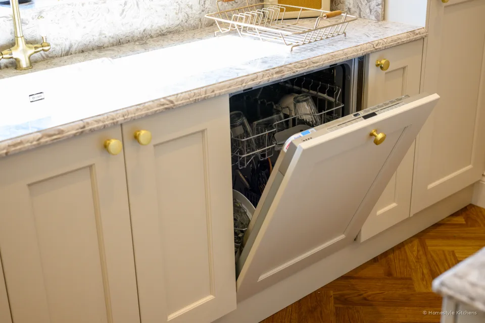 Integrated dishwasher open beside a cream sink run, marbled quartz and brass tap.