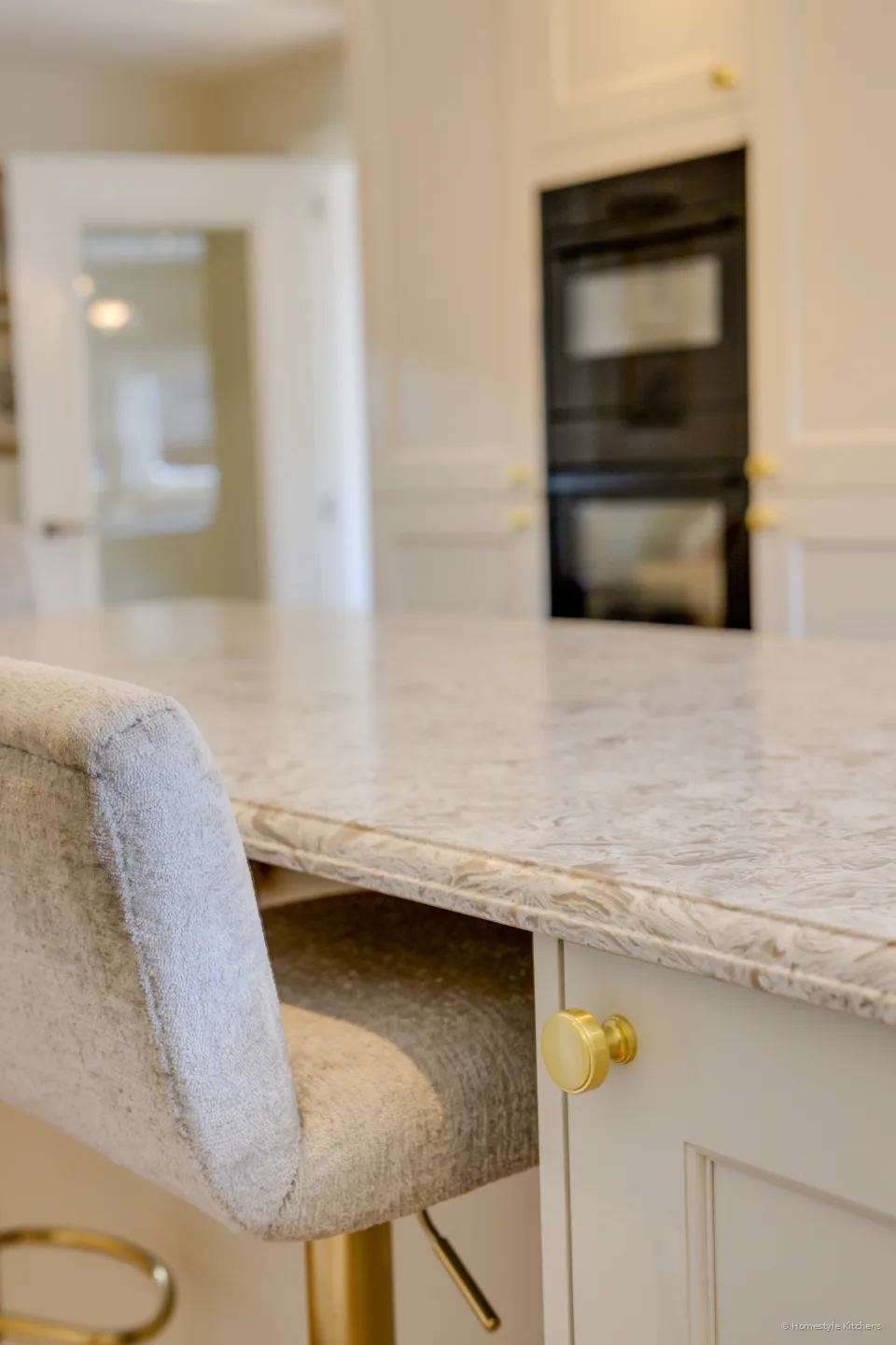 Low view along a quartz worktop toward black ovens in a cream tall housing.
