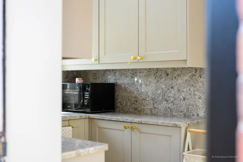 Cream wall cabinets with brass knobs, marbled quartz splashback and a black microwave on the worktop.