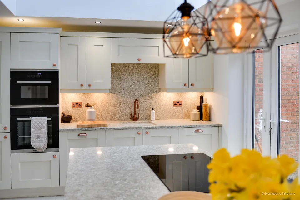 Ivory shaker oven housing with two stacked black ovens, copper knobs, quartz splashback and pendant lights.