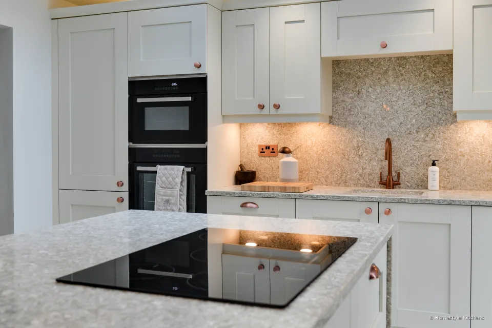 Black induction hob in a speckled quartz island, stacked ovens and copper knobs on the ivory shaker run behind.