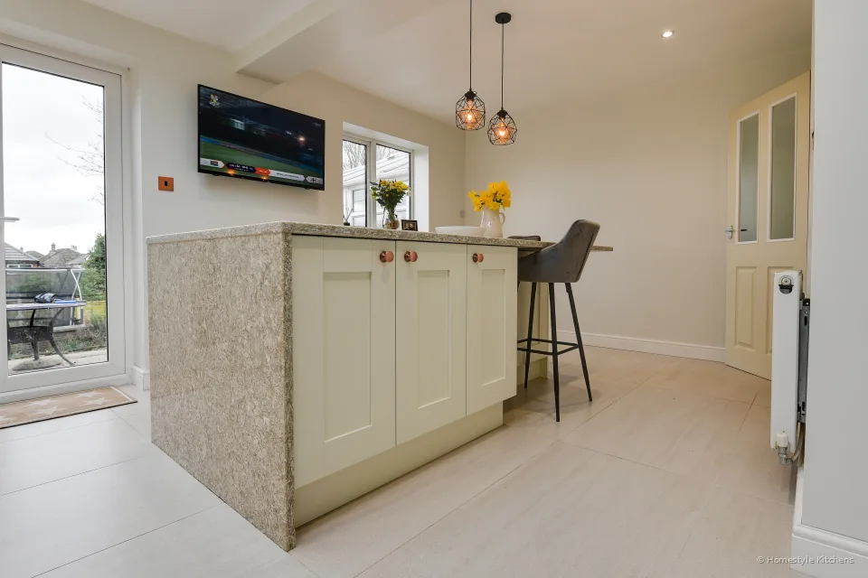 Ivory shaker island with a quartz waterfall end, grey stool, patio doors and a wall-mounted TV.