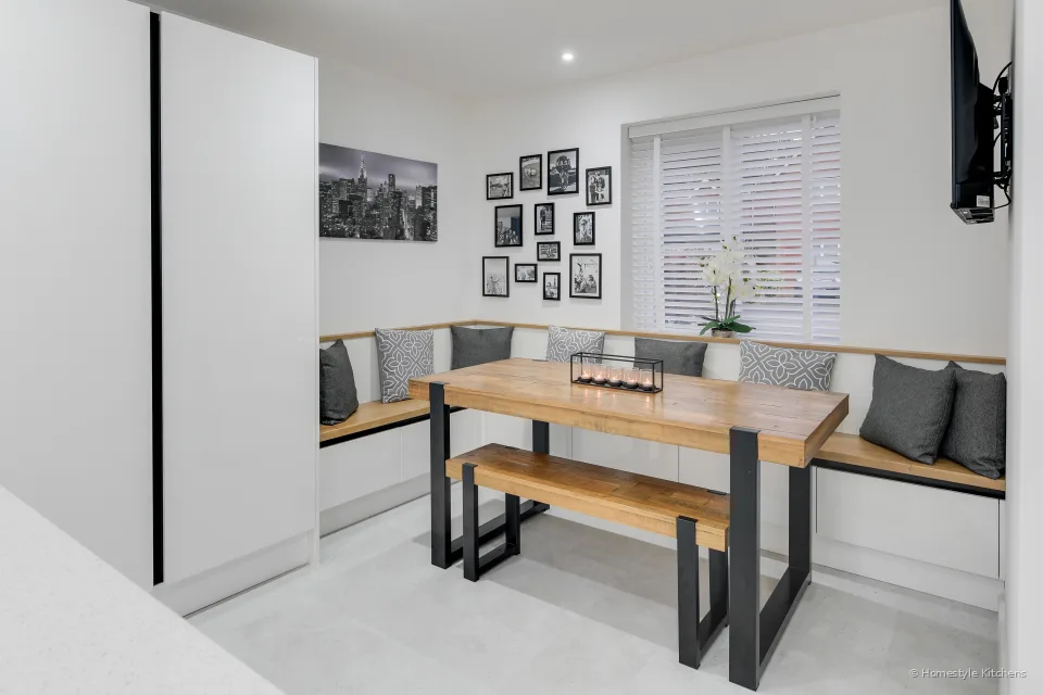 Built-in dining nook with an oak table and bench, grey cushions, gallery wall, tall white handleless units to the left.