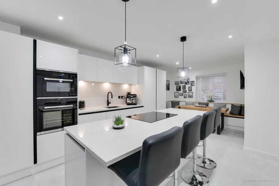 White gloss handleless kitchen, quartz island with a black induction hob and black bar stools, main run of units behind.