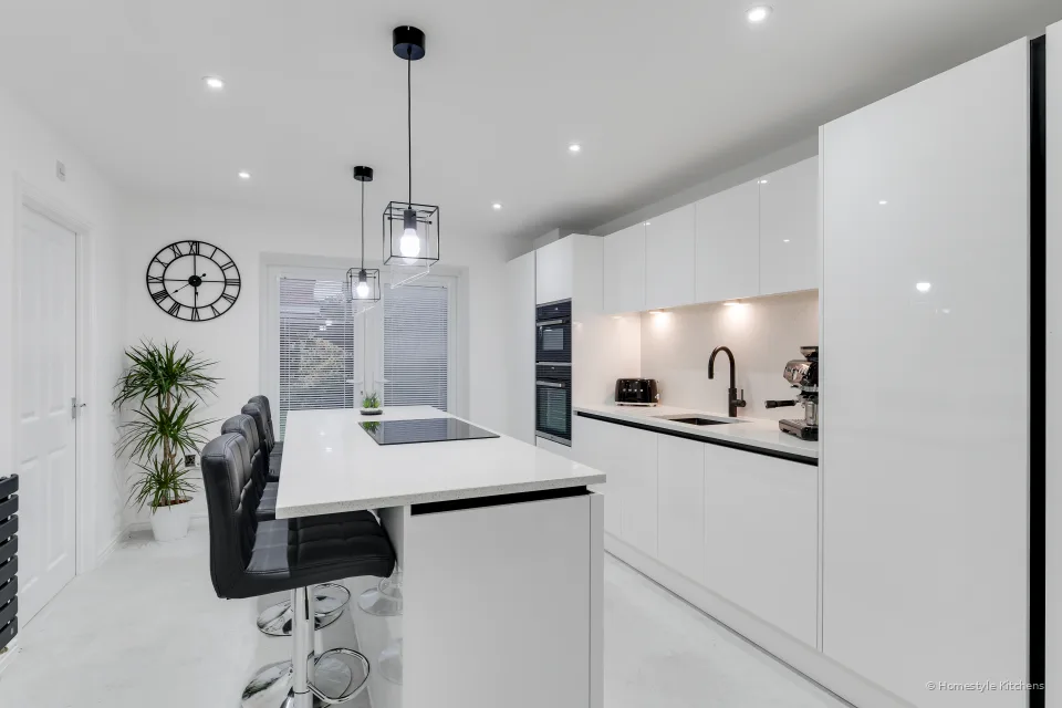 White gloss kitchen from the doorway, island with hob and bar stools, rear run with a black tap, wall clock and plant to the left.