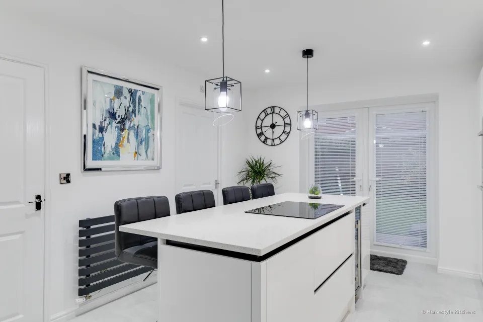 White quartz island with a black induction hob and bar stools, looking toward French doors, a wall clock and a plant.