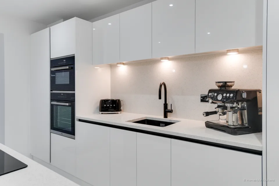 Rear run of white gloss units, under-cabinet lighting on a white quartz splashback, black tap and sink, oven and combi microwave to the left.