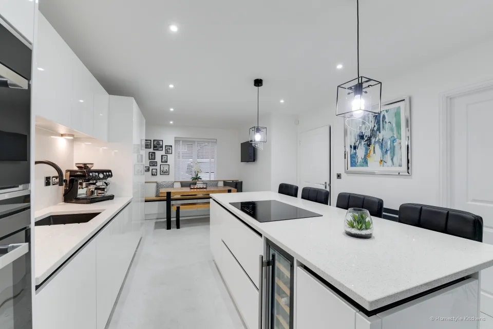 White handleless kitchen looking down the room, island with a wine cooler and black bar stools, dining nook at the far end.