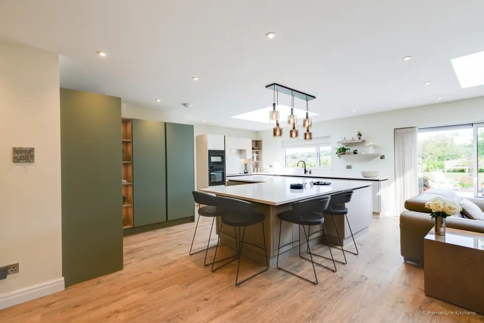 Open-plan handleless kitchen, cream stone units, sage talls, large island and copper pendants.