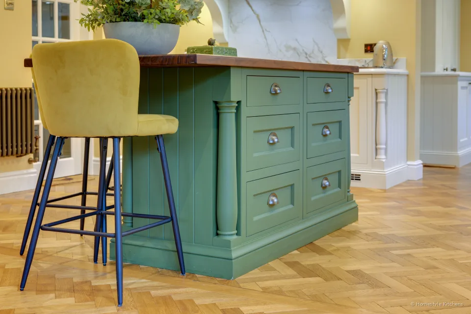 Corner of a green island with oak top, brass cup handles and a mustard bar stool on herringbone floor.