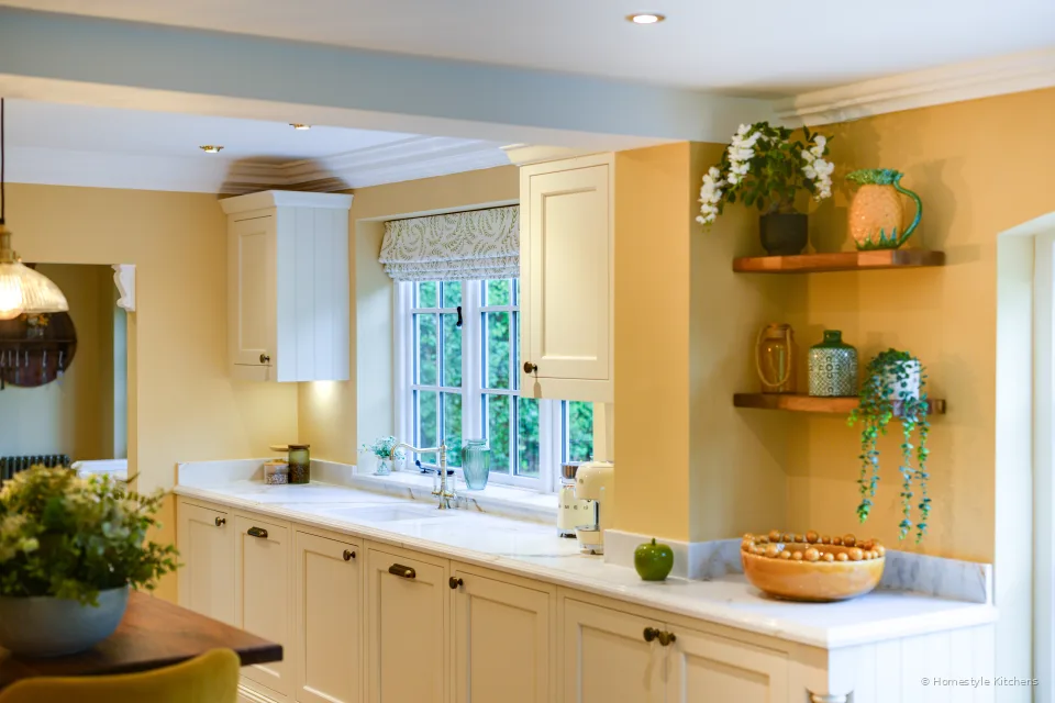 Cream painted kitchen run under a window with Calacatta Gold worktop, brass tap, and oak shelves.