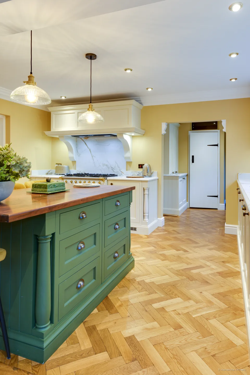 Portrait view past a green island toward a cream painted range wall and canopy.