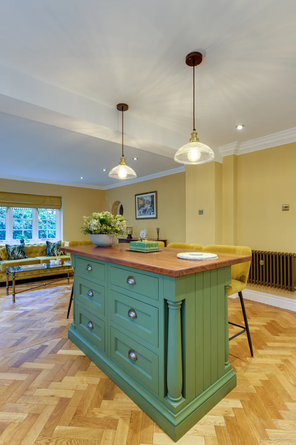 End of a green island with oak top and brass cup handles, pendants above, seating beyond.