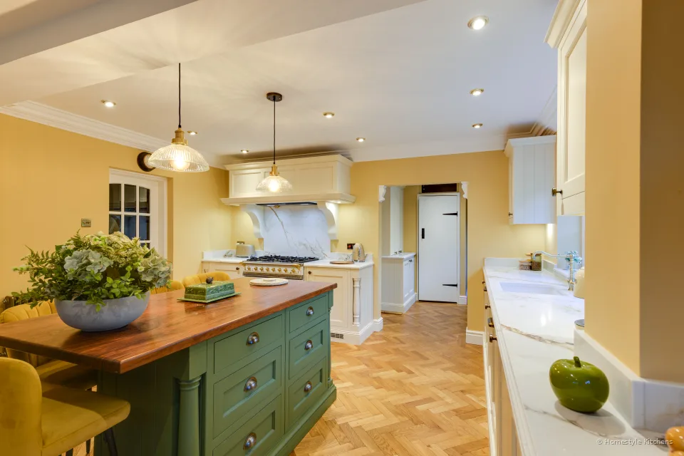 Wide kitchen with cream painted cabinets, a green island with an oak top, herringbone floor and a range under a canopy.