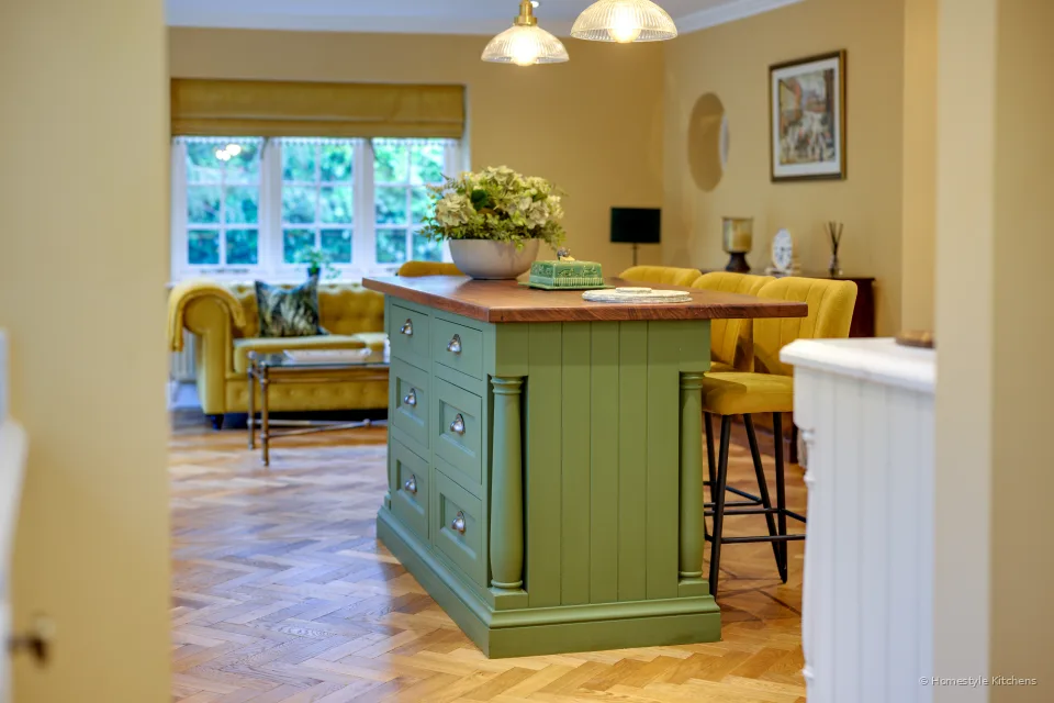 Green island with oak top and mustard stools, looking toward the sitting area and windows.