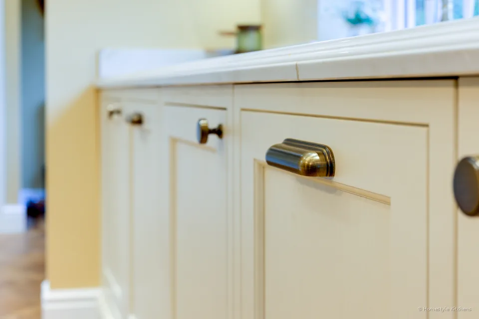 Close-up of a brass cup handle on cream painted in-frame cabinet doors under a quartz worktop.