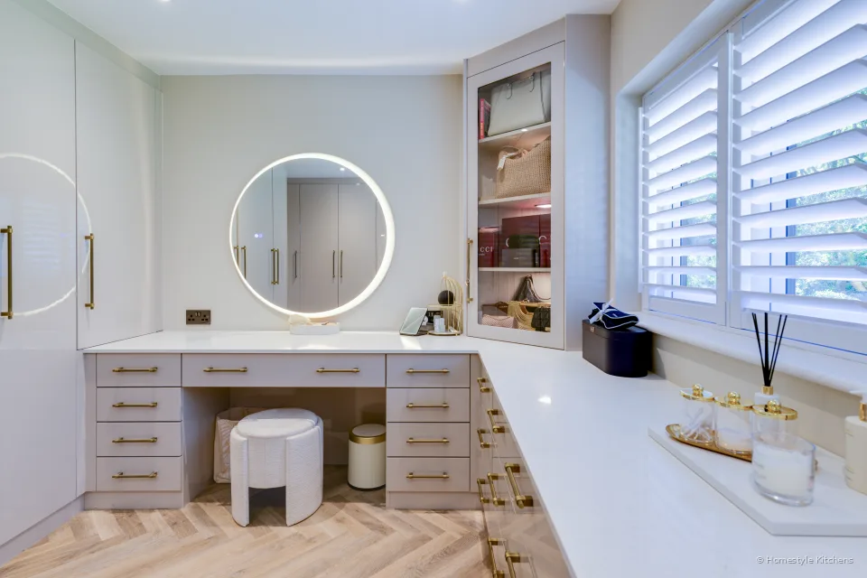 Pale gloss dressing table with a round lit mirror and drawers under shuttered windows.