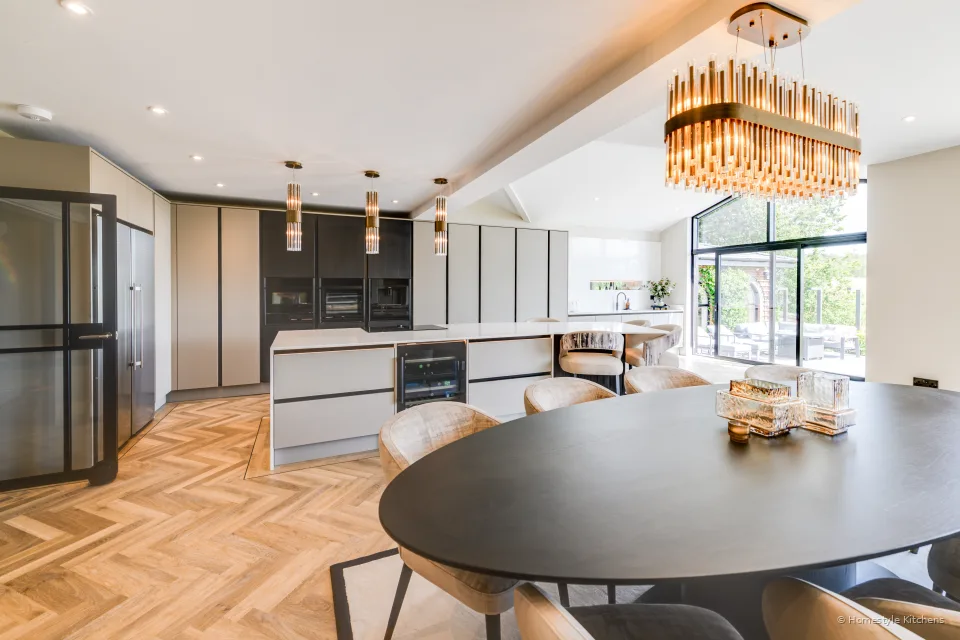 Pale grey kitchen island and a round dining table under brass pendants, herringbone oak floor.