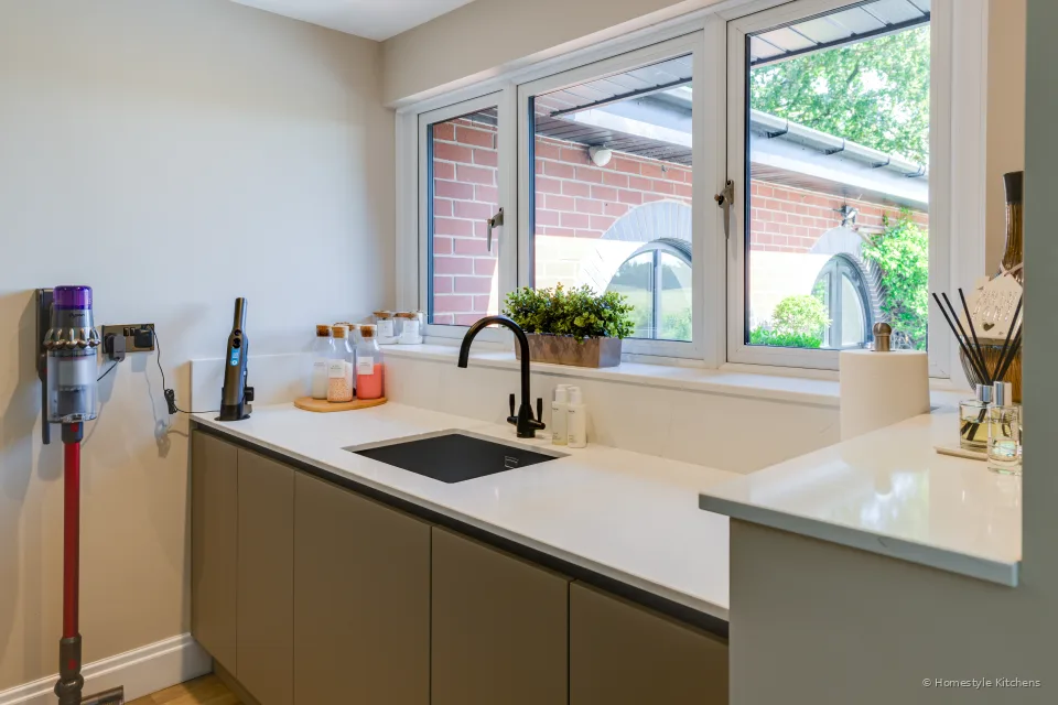 Taupe handleless utility, white worktop, black tap and undermount sink under a window.